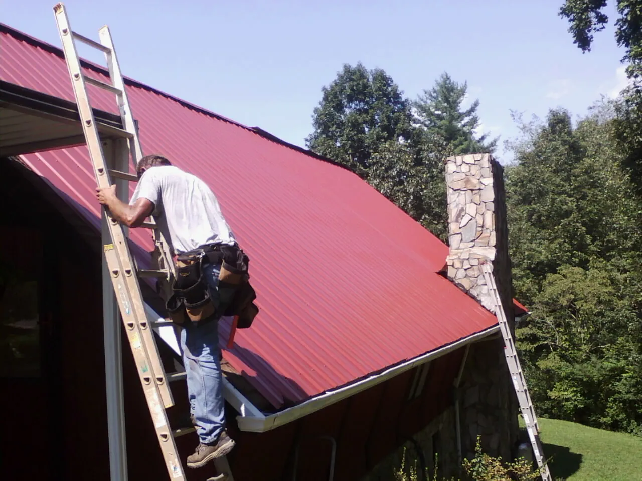 Roofer on a ladder beside a red metal roof and stone chimney — Soffit & Fascia Repair by Roofing Brothers in Tippecanoe